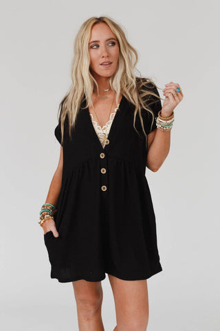 A woman with long, wavy blonde hair wearing the Sage and Wildflowers Romper - Black from Three Bird Nest, accessorized with multiple bracelets and rings, embodies a boho-chic style standing against a plain white background. She is looking slightly to the side with one hand raised near her face.