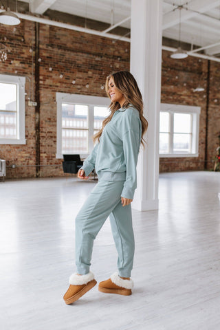 A person with long hair is wearing a relaxed fit Alison Half Zip Loungewear Set in light blue from Liam & Company and brown fur-lined slippers. They are standing in a spacious room with large windows, white wooden floors, and exposed brick walls, looking to the right and smiling.