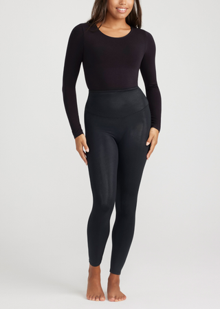 A person stands barefoot against a plain white background wearing a fitted, long-sleeve black top and Yummie's Elektra Coated Shaping Legging in black. They smile softly with their arms relaxed by their sides. The outfit showcases a sleek, all-black look while offering excellent tummy control with its seamless construction.