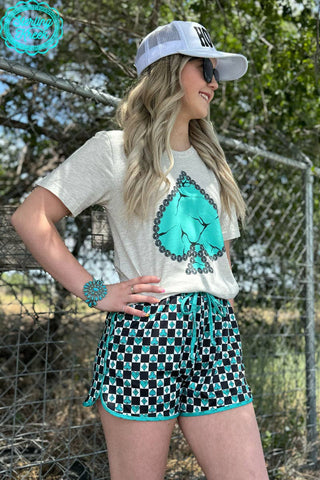 A person stands outdoors wearing a white hat, the Sterling Kreek My Lucky Day Tee featuring a decorative ace of spades design, and black shorts with a blue geometric pattern. They have long blonde hair accessorized with concho studs and a blue bracelet. A chain-link fence and trees are in the background.