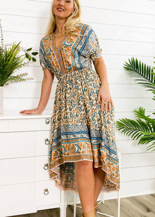 A woman in an Anna Kaci Wholesale Finn Tie Back High Low Dress, featuring a vibrant multicolored pattern, stands next to a white chest of drawers. She smiles slightly as one hand rests on the dresser. The background boasts white paneled walls and an array of green plants.