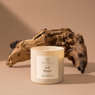 The 12oz "Rich Hippie" candle by Hico Candle Co. spreads its enchanting aroma, resting on a beige surface with driftwood nearby. The minimalist and earthy ambiance echoes the gentle bloom of a Flower Bomb in tranquility.