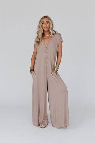 A woman with long blonde hair is wearing the Kendrick Mocha Jumpsuit from Three Bird Nest. The loose beige jumpsuit features short sleeves, a flattering V-neckline, and buttons down the front. She has several bracelets on both wrists and is standing against a plain white background, with her hands in the jumpsuit's pockets.