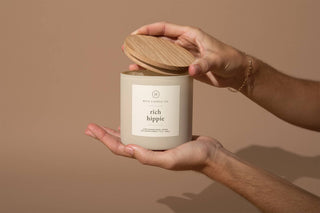 Two hands hold a 12oz candle with a wooden lid, labeled "Hico Candle Co." and "Rich Hippie," releasing scents of amber, orange, clove, and balsam. Set against a neutral beige background, this creation exudes elegant simplicity.