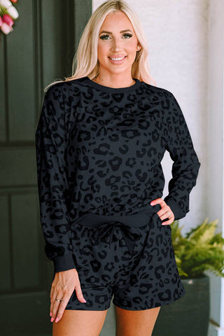 A person with long blonde hair stands in front of a dark-colored door, smiling. They are wearing the stylish and comfortable Wells 2 Piece Leopard Loungeseat by Lovesoft, made from lightweight material, in black and dark gray. Their left hand is on their hip, and there's a small green plant to the right.