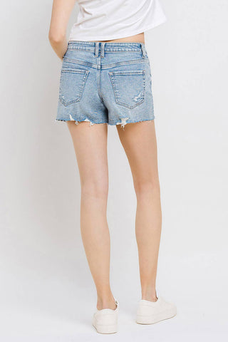 A person stands facing away, wearing VERVET by FLYING MONKEY Kylie Mid Rise Raw Distressed Hem shorts available in regular and curvy sizes, featuring slight distressing on the back pockets. They are also wearing a white top and white sneakers against a plain light-colored background.