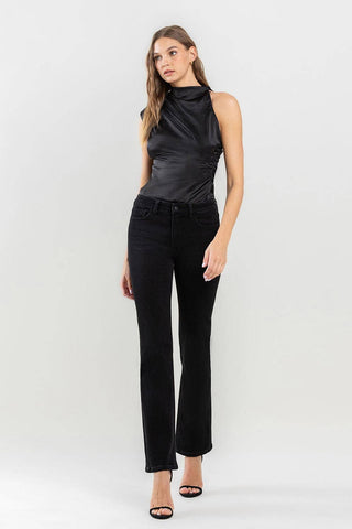 A woman with long hair stands against a plain background. She is wearing a sleeveless black satin top, Lovervet by VERVET's Halston Mid Rise Bootcut Jeans—an absolute wardrobe must-have—and black open-toe heels. She poses with one hand relaxed by her side and the other slightly bent.