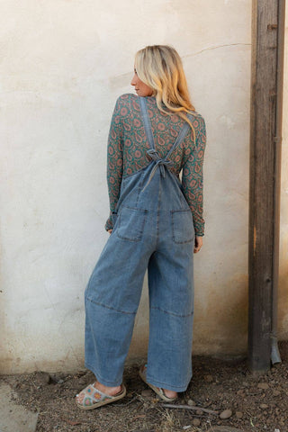 A person with long, blonde hair is facing away from the camera, dressed in the Dock Town Button Up Jumpsuit in denim by Three Bird Nest, paired with a patterned long-sleeve shirt that captures a boho style aesthetic. They stand against a weathered, light-colored wall next to a wooden post, with dirt and pebbles scattered on the ground beneath them.