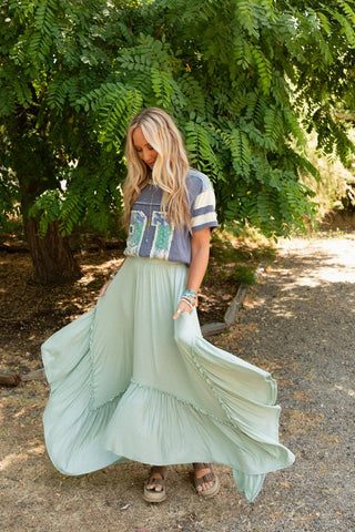 A woman with long, wavy hair stands outdoors on a dirt path, surrounded by lush greenery. She is wearing a loose, grey graphic T-shirt and the Logan Forever Beauty Maxi Skirt in Pale Sage from Three Bird Nest made of lightweight stretch knit fabric with a high-rise elastic waistband. She holds the sides of her skirt, lifting it slightly. Sandals and bracelets complete her look.