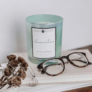 A lit "Illumination Candle" by Jewel Within, made from natural coconut wax and scented with "Valencia Orange," sits on a book. Resting on the book are a pair of tortoiseshell glasses, dried flowers, and a 925 Sterling jewelry crescent moon necklace. The plain white background creates a calm and minimalist setting.