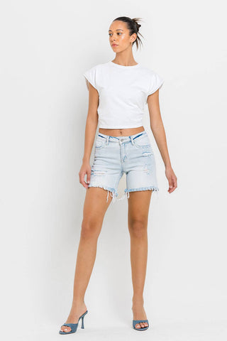 A woman stands against a white background wearing a white short-sleeve crop top, the Kolson Mid Rise Distressed Stretch Denim Shorts by VERVET by FLYING MONKEY with a frayed hem, and blue high-heeled sandals. Her hair is styled in a high ponytail.