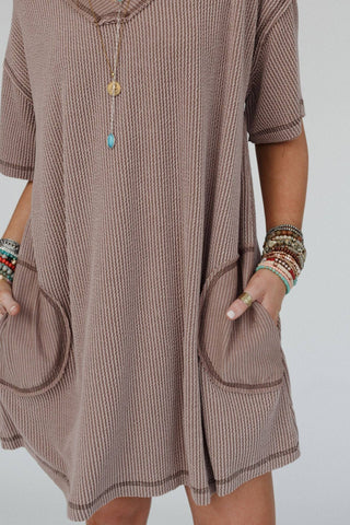 A person wearing the Brooklyn Soft Touch Tunic Dress by Three Bird Nest, a loose, short-sleeved boho-chic design with pockets, stands with their hands in the pockets. They accessorize with colorful beaded bracelets, various rings, and layered necklaces featuring pendant charms. The comfortable stretch fabric of the dress complements the plain, light-colored background.