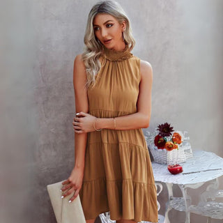 A woman with long, wavy blonde hair stands beside a white table, holding a beige clutch. She is dressed in a sleeveless Scottie Halter Summer Dress by MISS SPARKLING, featuring a camel color, ruffled high neckline, and lightweight design. To complement her outfit, she wears turquoise jewelry. On the table rests a flower arrangement and a red apple against the backdrop of a neutral wall.