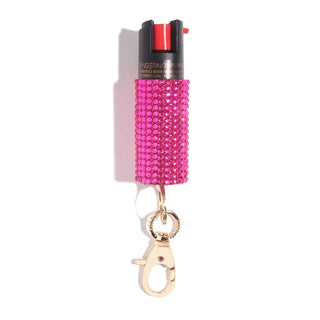 A small container covered in pink rhinestones, featuring a gold keychain clip at the bottom, designed by BLINGSTING, a women's safety brand. The Emergency Pepper Spray Key Ring has a black top with a red button and includes UV marking dye for added security.