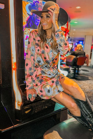 A woman in the Hollan Concho Grooves Dress by 2 Fly and a black cowboy hat sits on a slot machine in a casino. She is adjusting her hat with one hand, smiling, and wearing black cowboy boots with turquoise snaps. The background features more slot machines and patrons sitting at them.