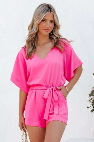 A woman with wavy blonde hair is wearing the Emmitt V-Neck Tie Waist Short Romper in bright pink from Charmo. She stands against a plain white background and holds a gold chain in her right hand, while her left hand rests on her hip.