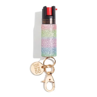 Introducing the Emergency Pepper Spray Key Ring by BLINGSTING: a glittery, colorful striped canister with a keychain attachment and a gold-tone clasp. The keychain features a round, gold tag inscribed with "BLINGSTING." Designed to enhance women's safety, the canister includes a black top with a red activation button and UV marking dye for added security.