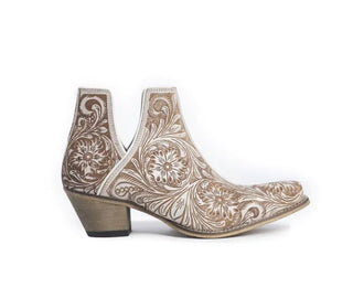 The White Chocolate Tooled Booties by Myra feature a tan ankle boot adorned with intricate white floral and swirling patterns carved into its surface. These Western hand-tooled leather booties showcase a low, thick heel with a wooden texture, blending traditional and contemporary styles seamlessly in an ornate and artistic design.