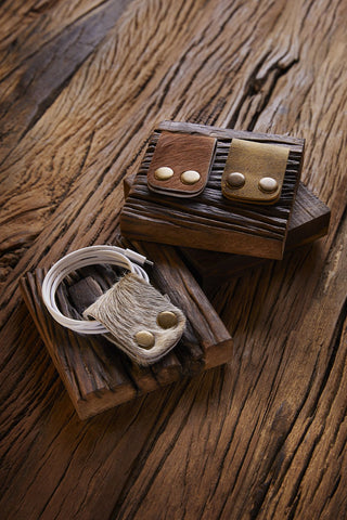 A wooden surface holds rustic stands displaying leather accessories. One stand features two brown and tan leather card wallets with button fasteners, while the other showcases the Myra Daylynn Cable Organizer, with charging cables neatly coiled in a fur-textured leather holder secured by two buttons.