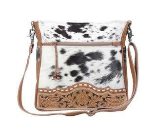 Introducing the Myra Cowhide Hand Tooled Leather Handbag, Western Handbag: This stylish crossbody bag showcases a striking black and white cowhide design complemented by a brown leather trim. It features a front zipper pocket, an adjustable strap, and embossed leather trim adorned with an intricate floral and leaf pattern.