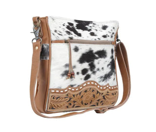 The Cowhide Hand Tooled Leather Handbag by Myra is a stylish Western handbag featuring a leather strap and floral tooled details at the bottom. This elegant bag includes a front zipper pocket, an embossed leather rug design, and showcases a black-and-white spotted fur pattern on the main body.