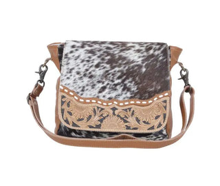 The Mangnifique Tooled Hide Hair Handbag-Concealed Bag by Myra is a stylish crossbody accessory featuring a chic black and white hair-on cowhide flap accented with brown leather trim. The intricately detailed tan floral embroidery on the flap adds elegance, while the adjustable brown leather strap with metal clasps and zippered compartments provide added convenience.