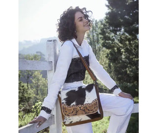 A person with curly hair is leaning against a white wooden fence, smiling with their eyes closed. They are wearing a white top and light-colored pants, and carrying the Mangnifique Tooled Hide Hair Handbag-Concealed Bag by Myra, which is black and white with intricate brown detailing. Trees are visible in the background.