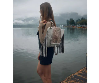 A person wearing a light brown long-sleeve top and dark overalls stands on a dock by a foggy lake, carrying the Myra SMOKEY BACKPACK Leather & Hairon Bag. They face sideways, looking ahead with mountains and trees visible in the background.