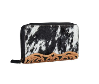 The Darrington Ranch Cowhide Tooled Wallet by Myra is a black and white hair-on hide wallet adorned with intricate brown leather trim featuring a floral design. This distinctive cowhide accessory includes a zippered closure and a hand-tooled leather zip pull tab.