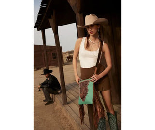 A woman dressed in Western attire, including a cowboy hat, white sleeveless top, and brown shorts, stands in front of a wooden building with a porch. She holds the Myra Terra Concealed Carry Handbag Tooled Leather in turquoise and wears turquoise cowboy boots. A seated man in similar attire is visible in the background.