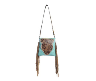 The Terra Concealed Carry Handbag Tooled Leather by Myra is a turquoise leather bag with a removable shoulder strap, featuring an ornate, brown embossed leather flap and long brown fringe details hanging from both sides. The hand-tooled detailing on this design beautifully combines Western and bohemian elements.