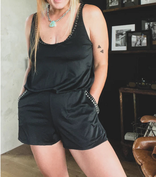 A person is dressed in a black Rowdy Mama Swim Romper from 2 Fly, with their hands tucked into the pockets. They accessorize with a turquoise necklace and showcase a tattoo of butterflies on their right arm. The background features a wall adorned with framed photos and a wooden shelf displaying an assortment of items, including some apparel from Ultimate Mom Swim.