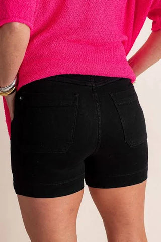 A person wearing a bright pink top and Hollis Black Button Fly High Rise Shorts Regular Curvy Judy Blue Shorts stands with their hands resting on their hips. The focus is on the lower back and shorts, showcasing the non-distressed style of this Judy Blue casual outfit.