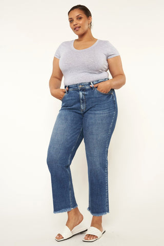 A woman stands confidently against a plain background, wearing a light gray fitted T-shirt and the Kan Can USA Kimber Curvy High Rise Slim Straight Jean in medium stone wash with frayed hems. She has her hands in her pockets and completes her look with white slide sandals.