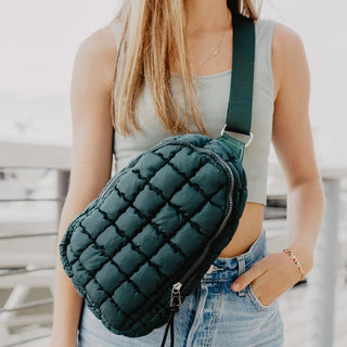 A person with long hair, wearing a light green tank top and blue jeans, carries an Ellison Quilted Crossbody Handbag from Western Edge Boutique with an adjustable strap across their chest. The background is blurred and appears to be an outdoor setting.