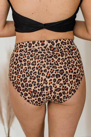 A person is sporting the Renley Shores High-Rise Swim Bottoms in Natural Leopard by MACK & MAI, paired with a black top. The image highlights the lower back and bottom half, emphasizing the bikini's 80% Nylon 20% Spandex material and its stretchy waistband. The backdrop is neutral with touches of dried plant decor.