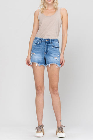 A person is wearing a light brown sleeveless top, VERVET by Flying Monkey's Harlow Super High Rise Stretch Mom Shorts with a raw hemline, and beige sneakers. They are standing against a plain white background. The outfit has a casual and relaxed style, perfect for showcasing the comfort of denim in everyday wear.