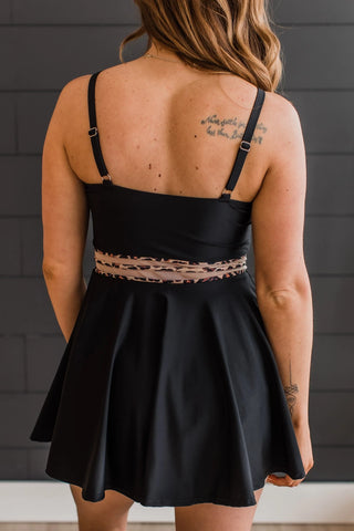 A rear view of an individual wearing the "Living On Sunshine Swim Dress" in black with leopard accents from Western Edge Boutique features a flared skirt and thin straps. The person, showcasing highlighted brown hair and visible tattoos on their back, stands against a dark wall.