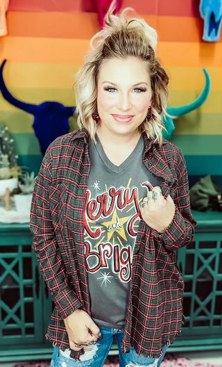 A woman with wavy blonde hair stands in front of a colorful rainbow wall, wearing the "Holly Jolly Flannel" by One24rags over a "Merry and Bright" t-shirt and ripped jeans. She smiles softly, surrounded by decor featuring bull head sculptures.