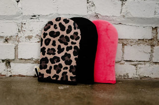 Three reversible tanning mitts are displayed against a white brick wall. From left to right, there is the Leopard Self Tanner Application Mitt by Western Edge Boutique, a black mitt, and a bright pink one. Perfect for applying tanning mousse, these mitts are standing upright on a concrete surface.