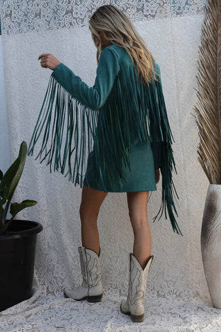 A person with long blonde hair, wearing the Flynn Suede Fringe Dress by Blue Buttercup, stands with their back to the camera. They are also wearing white cowboy boots and are positioned in front of a white lace backdrop with a tall plant and vase nearby.