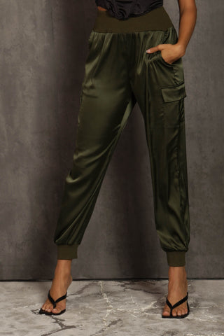 A person is wearing Blue Buttercup's Ora Olive Satin Cargo Pants with side pockets and black heeled sandals. The olive green satin fabric complements the textured gray wall and marble floor in the background. The person's upper body is not visible in the image.