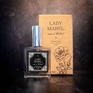 A bottle of Lady Mabel Perfume by R. Rebellion, a fine fragrance, is displayed against a dark background along with its brown packaging box that features a floral illustration. The bottle is adorned with a silver cap and an ornate black and white label. The name "Lady Mabel" is prominently visible on both the paraben-free bottle and the box.