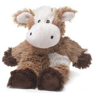 The Mills Moo Cow Junior Warmies from Warmies is a plush cow with soft, fluffy brown fur and a white snout, belly, and forehead. It features small white horns, floppy ears, and large black eyes. This microwaveable plush releases a soothing scent of French lavender and sits upright with a friendly and cuddly appearance.