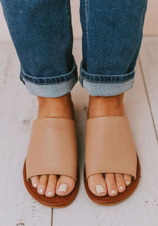 Close-up of a person wearing blue cuffed jeans and comfortable Quinn Nude Slide Sandals by Western Edge Boutique, standing on a light wooden floor. Their toenails are neatly manicured with a light nude polish. The sandals fit true to size, complementing the casual yet chic look.