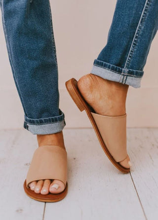 A person wearing cuffed blue jeans and tan Quinn Nude Slide Sandals by Western Edge Boutique. These comfortable sandals feature a wooden sole and an open-toe design, showcasing polished toenails. The person’s right foot is slightly raised, revealing the sandal’s flat heel—true to size against a light-colored floor background.