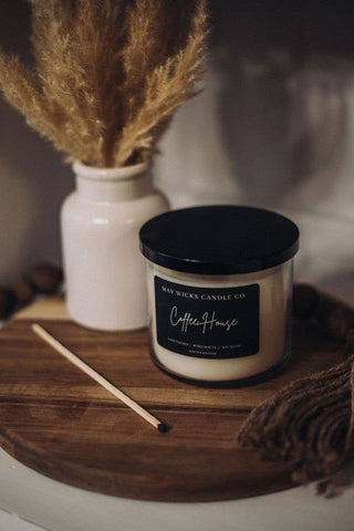 A white jar filled with dried wheat stalks is placed next to a Mahogany Teakwood Candle: 17 OZ from Mav Wicks Candle Co labeled "Coffee House" on a wooden tray. A long matchstick lies in front of the candle, and a piece of brown fabric is partially visible on the right side of the tray, hinting at its sophisticated fragrance.