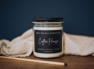 A glass jar candle labeled "Mav Wicks Candle Co - Luxe(Diva) Candle: 8 OZ," sits on a wooden surface, serving as the perfect home decor item. The background features a soft beige knitted fabric, and a matchstick rests nearby, enhancing the cozy ambiance of this best-selling fragrance.