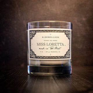 A clear glass candle with a white label sits against a dark background. The label reads "R. Rebellion Miss Loretta Candle" at the top, followed by "Made in the West" and "8 oz. 227 g. Pendleton, Or." The label features an ornate border design, hinting at its luxurious coconut soy wax blend.