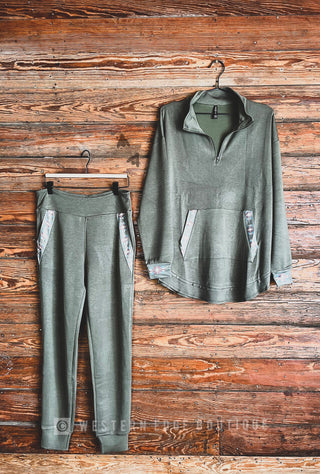 A soft olive green Scarlett Hot Trot Jogger Set from Western Edge Boutique hangs against a wooden background. The set features a long-sleeve quarter-zip pullover with Aztec detailed cuffs and matching pants, both adorned with subtle floral accents on the pockets. The label "Western Edge Boutique" is visible at the bottom of the image.